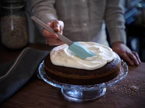 Pekař pomocí gumové stěrky nanáší krém na dort na skleněném servírovacím stojanu.