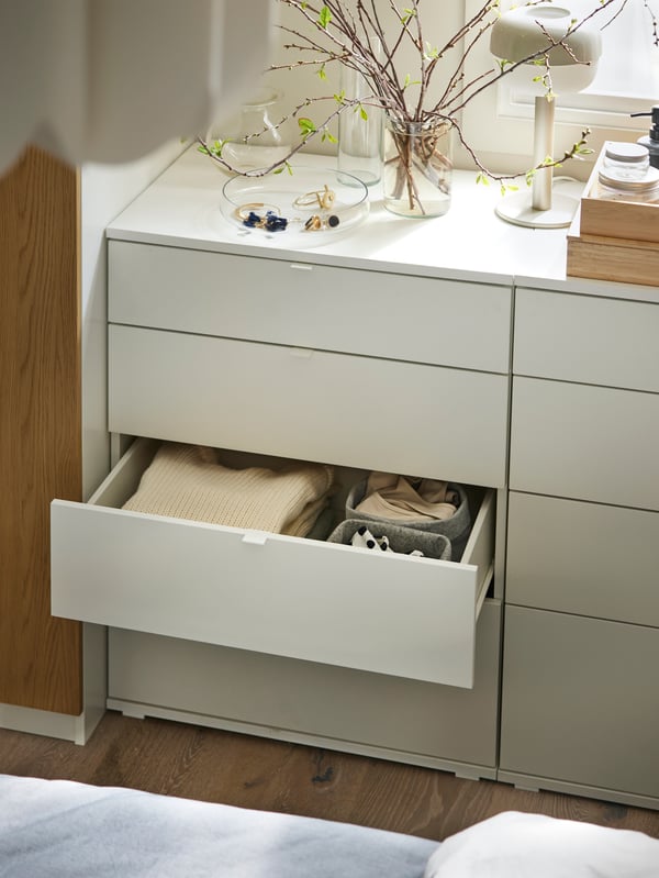 A white VIHALS chest with 4 drawers with the middle drawer open and a beige BLÅSVERK table lamp and vases on the top.
