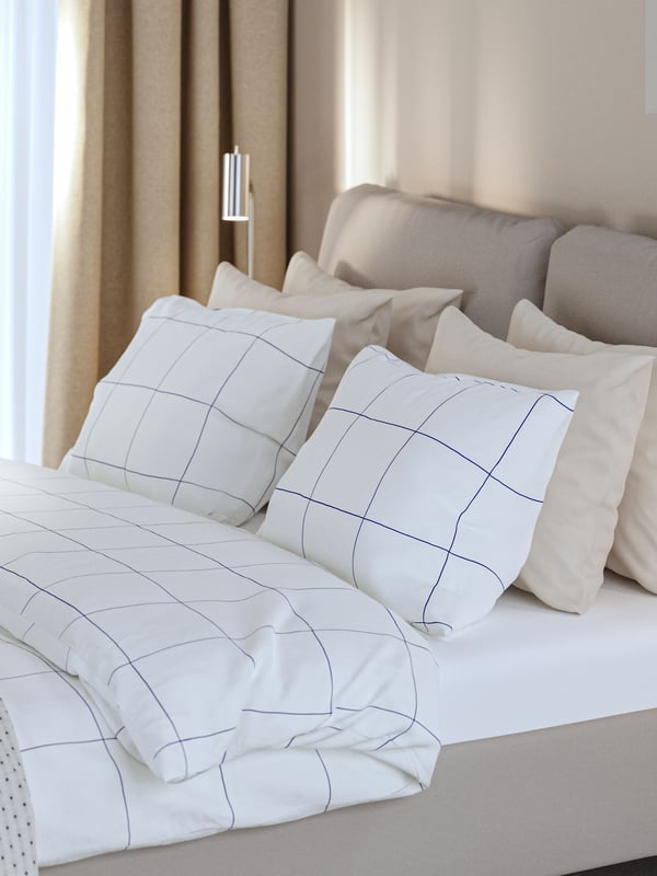 A hotel room with a SAGESUND upholstered bed made with BREDVECKLARE bedding next to a bedside table in oak veneer.