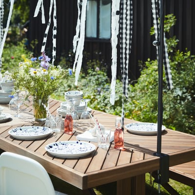 La taula de fusta NORRMANSÖ del jardí està preparada i decorada per a una celebració d'estiu.
