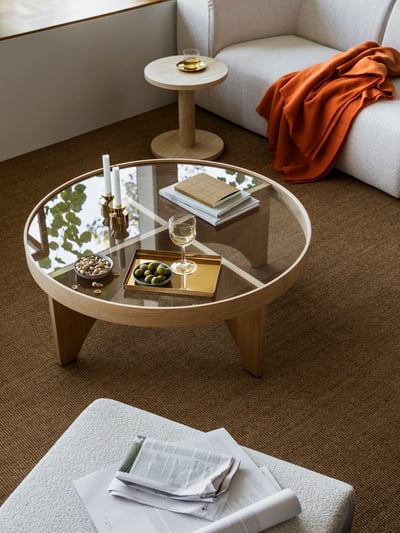 A brass-coloured tray on a STOCKHOLM 2025 coffee table in oak veneer and glass placed next to a pine veneer side table.