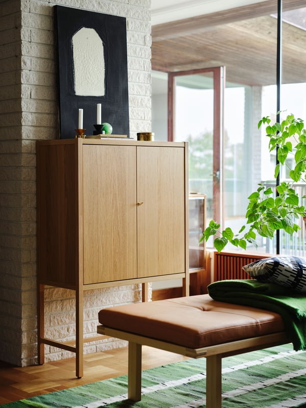 An oak veneer STOCKHOLM 2025 cabinet with two doors against a white brick wall, with a bench with a leather cushion in front.