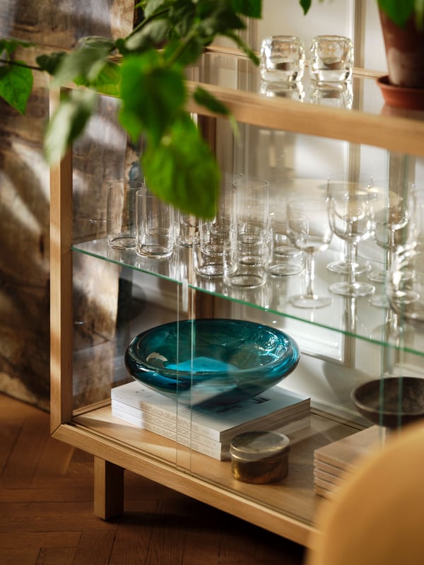 A dark blue-turquoise STOCKHOLM 2025 bowl on a stack of books, placed on a shelf in an oak veneer glass door cabinet.