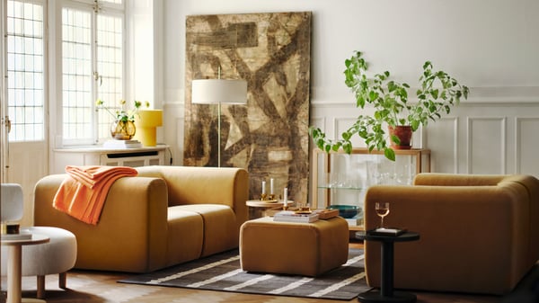 A bright living room with two beige STOCKHOLM 2025 2-seat sofas facing each other, with a pouffe on a rug between them.