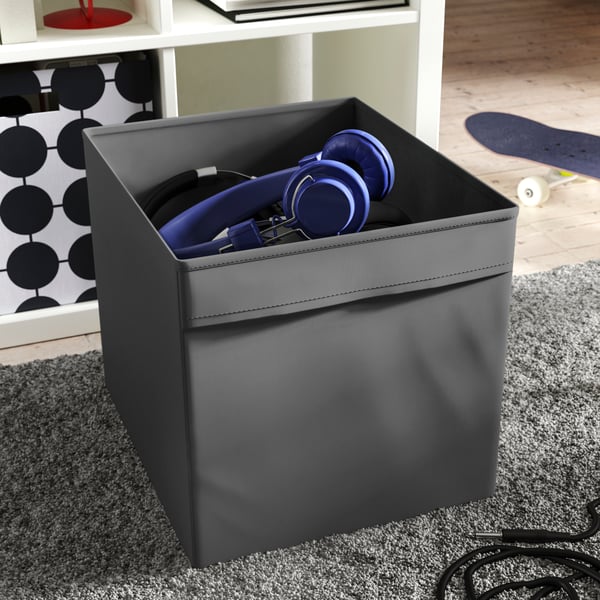 A dark-grey DRÖNA box holding pairs of headphones is placed on a high-pile rug beneath a white KALLAX shelving unit.